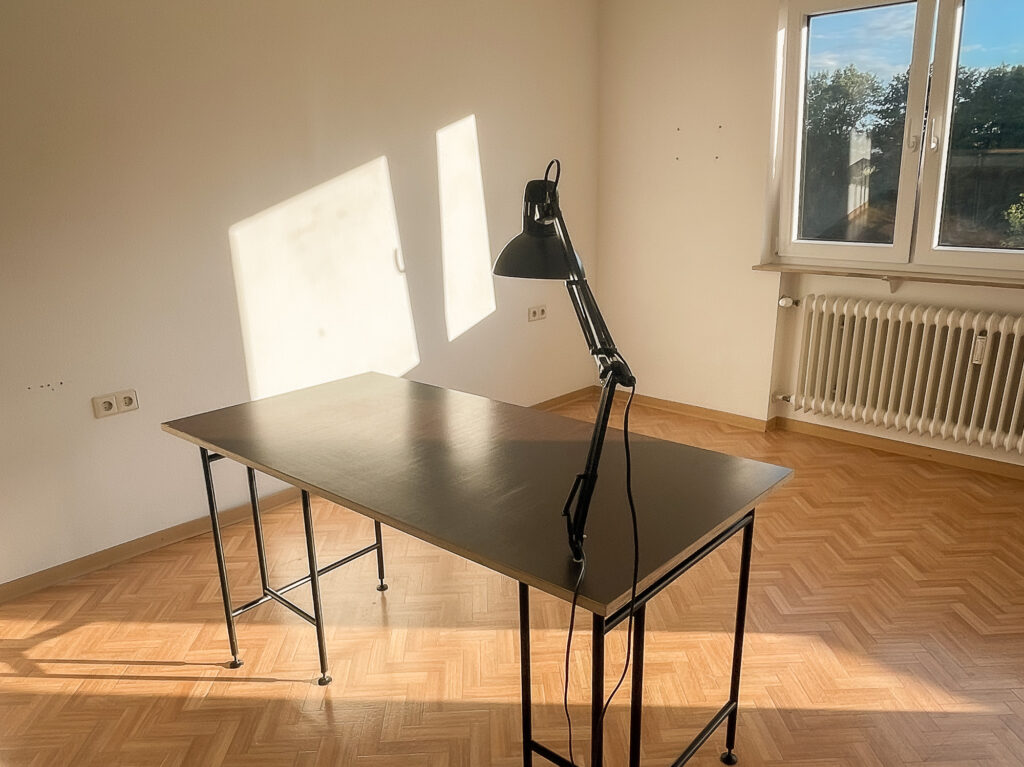 Ein einfacher Schreibtisch mit dunkelbrauner Multiplexplatte steht verloren in einem ansonsten leeren Zimmer mit weißen Wänden.